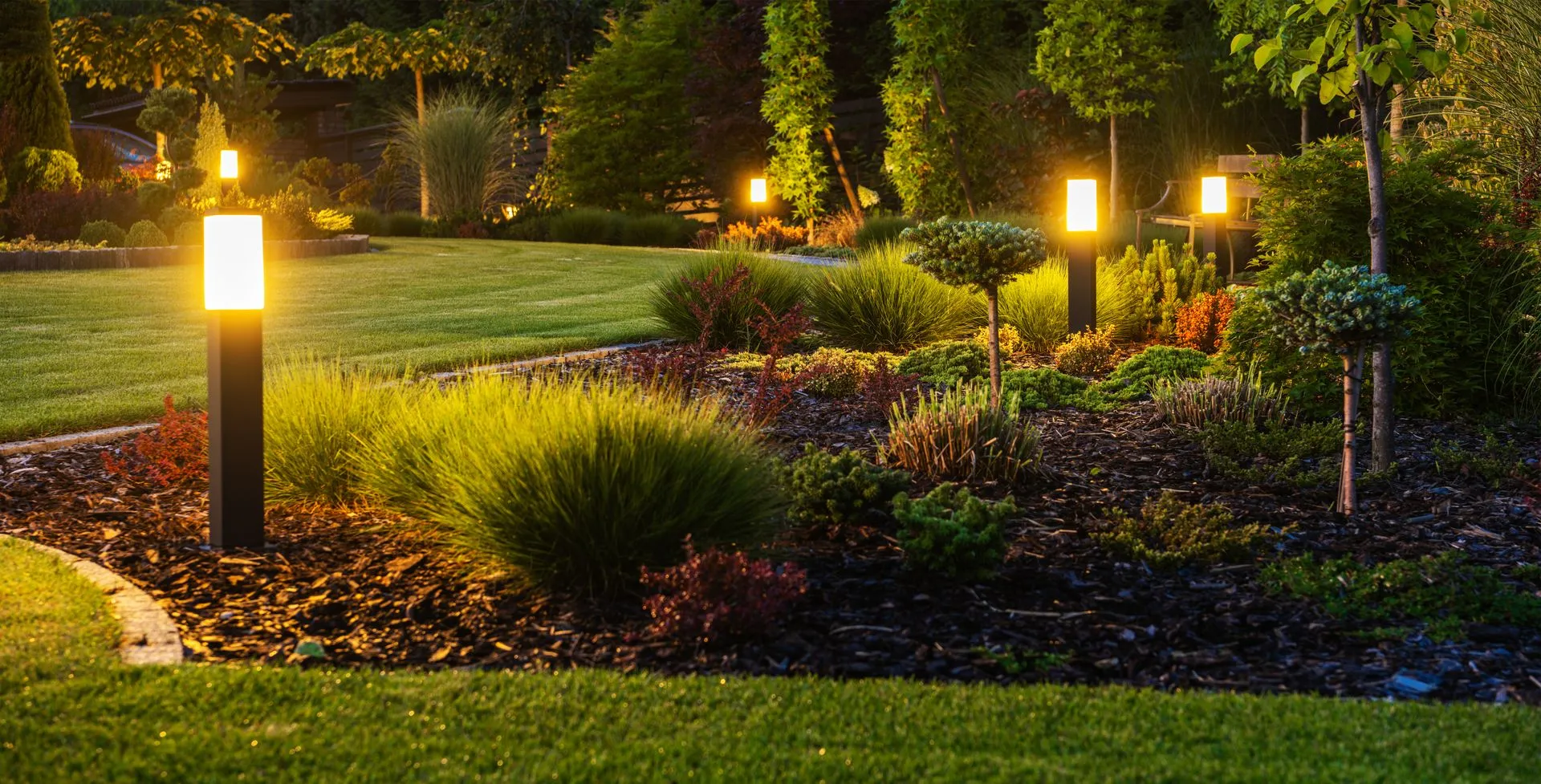 Lush garden at dusk with illuminated pathway lights, bushes, and green grass.