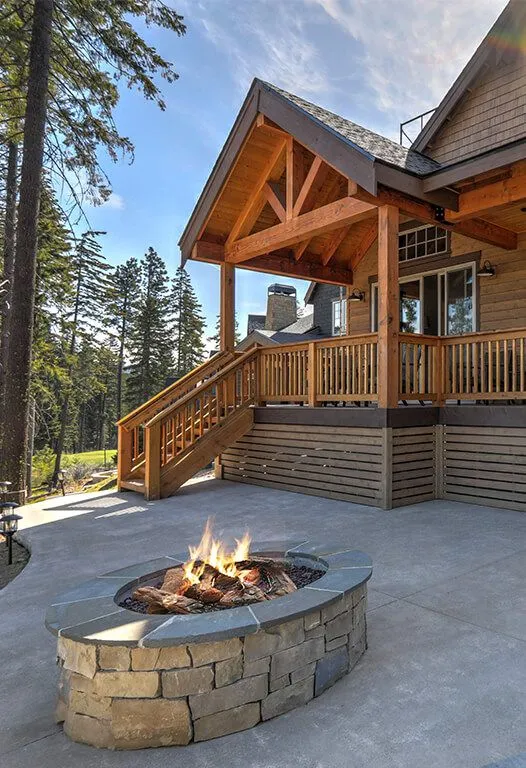 Exterior of a wooden house with a deck, steps, and a stone fire pit on a concrete patio, trees in background.
