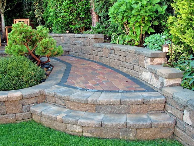Stone steps and curved wall enclose a brick patio in a garden with green plants and grass.