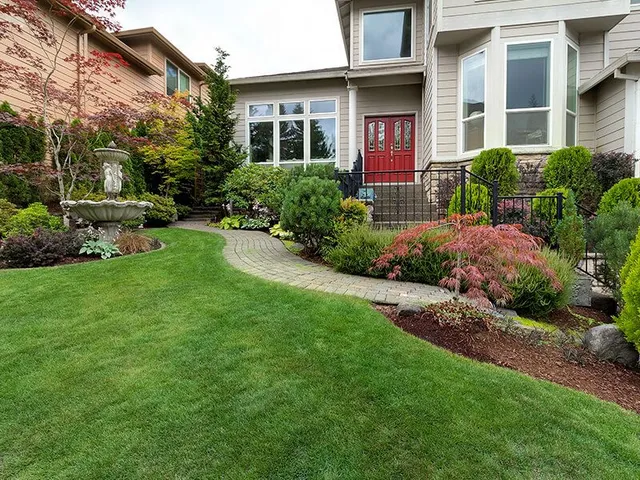 Well-manicured front yard with a winding brick path leading to a red front door. Lush green lawn and colorful shrubs.