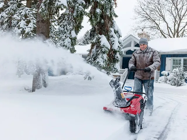 Man using snowblower to clear a snowy path in front of a blue house.