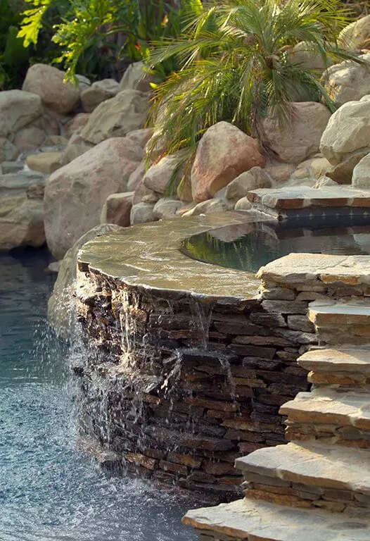 Waterfall cascading into a pool with stone steps, surrounded by rocks and greenery.