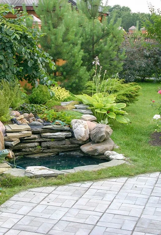 A garden with a small pond cascading over rocks, green plants, and a stone path.