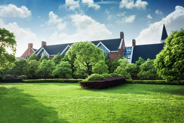 Lush green lawn in front of a brick building with a dark roof, surrounded by trees under a blue sky with clouds.