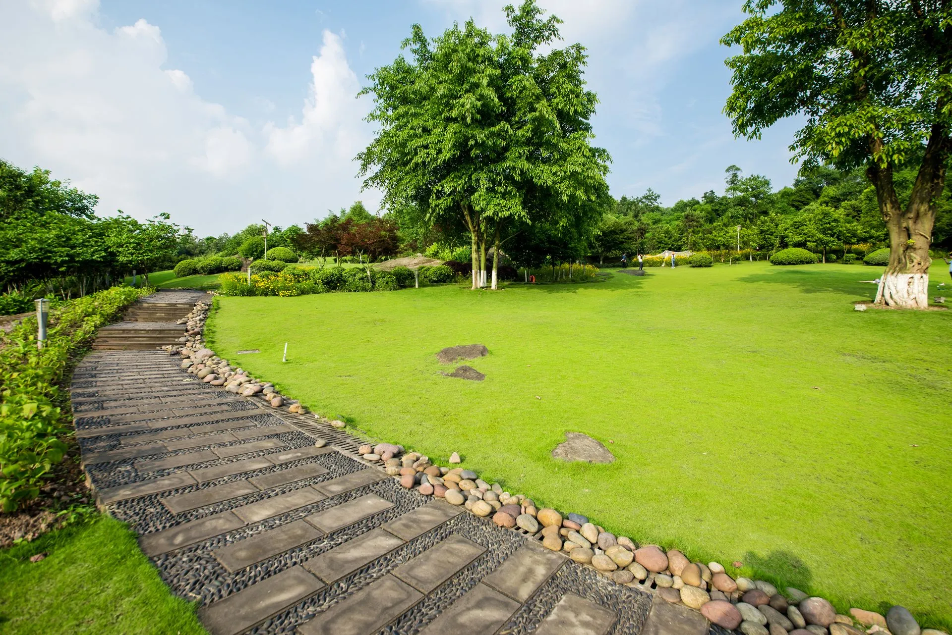 Stone path winds through a green lawn, past trees under a bright blue sky.