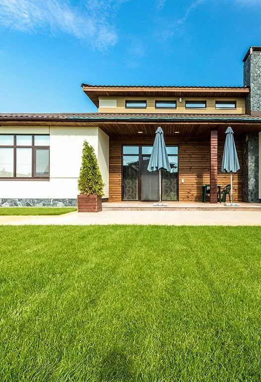 Green lawn leads to a modern house with wooden facade, patio, and blue umbrellas under a blue sky.
