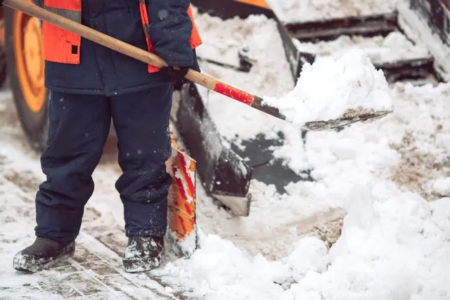 Person in blue coat and orange vest shoveling snow near a snowplow.